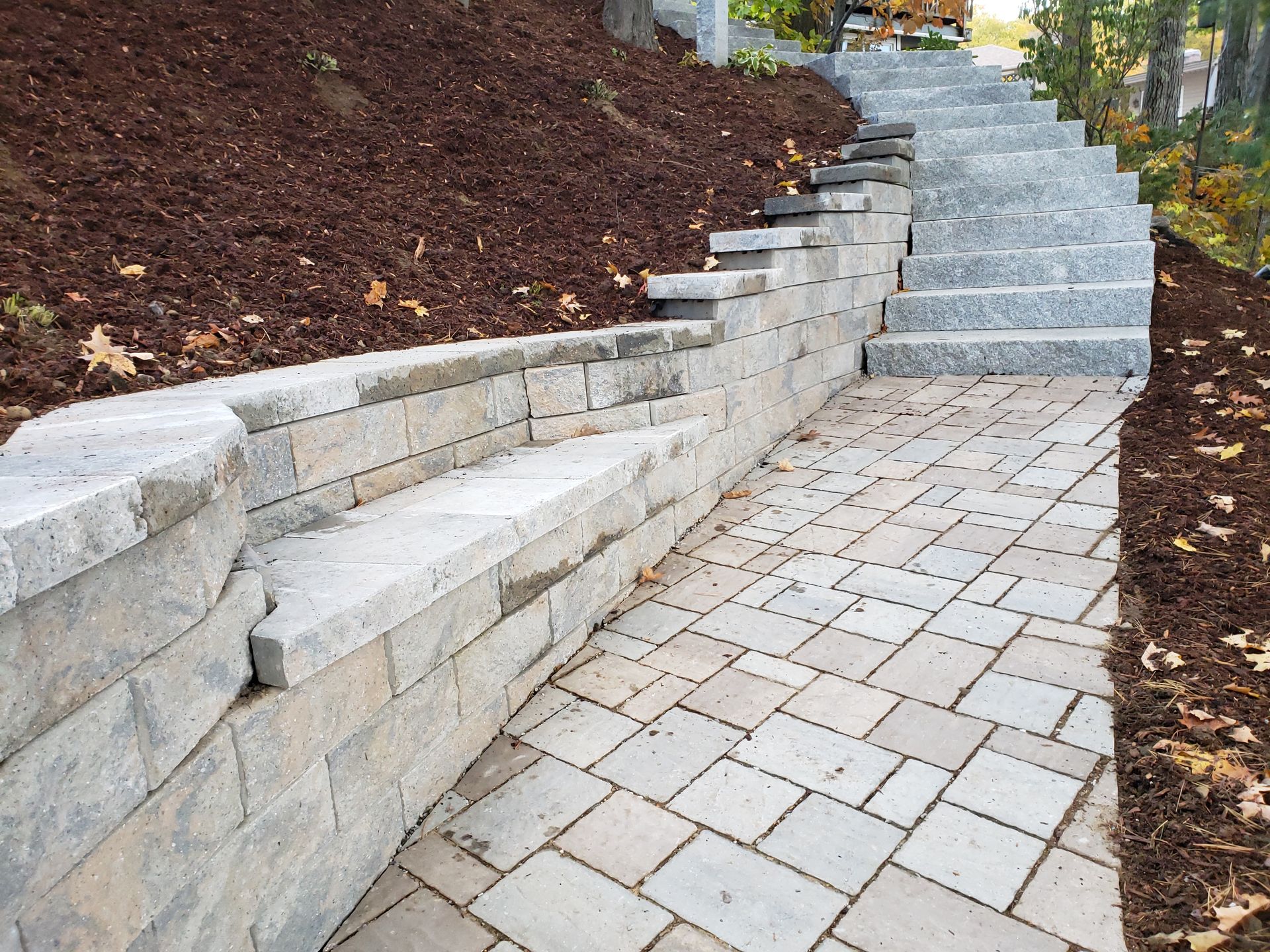 Stone retaining wall and staircase next to a paved path with mulch and trees.