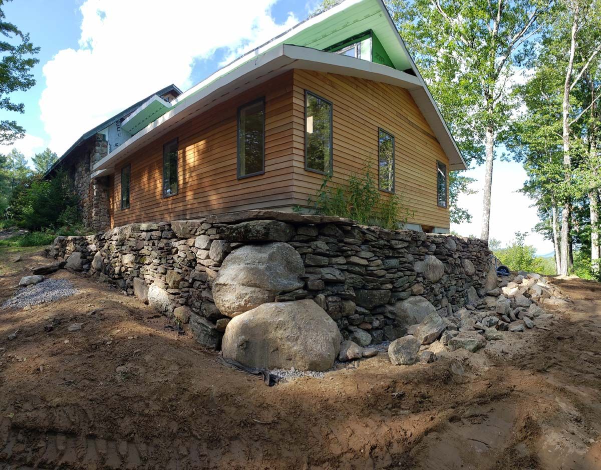 Stone wall supporting a wood-sided house. Light brown siding, green roof, windows. Dirt foreground, trees in background.