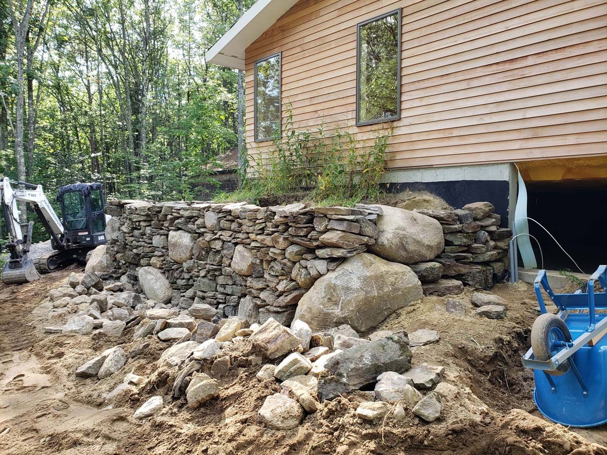 Stone retaining wall under construction next to a wood-sided building. A small excavator and wheelbarrow are nearby.