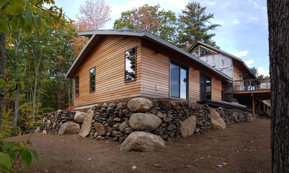 Wooden house under construction with stone wall, surrounded by fall trees and foliage.