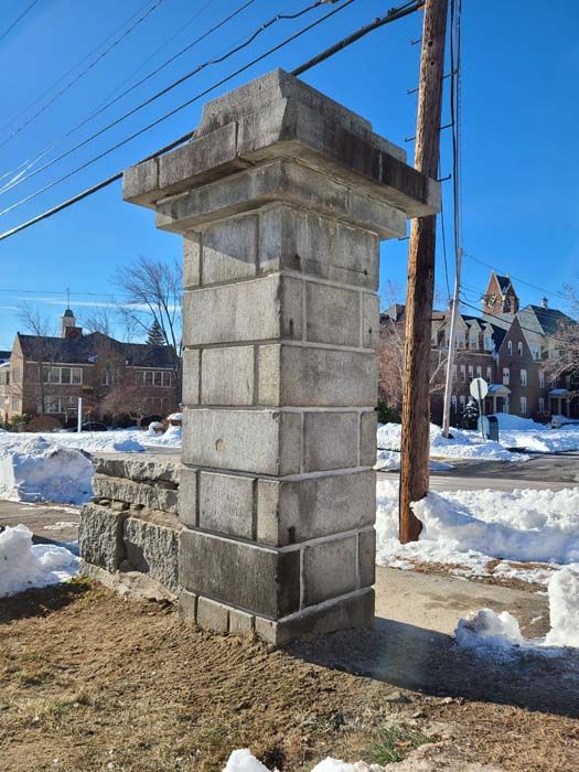 Stone pillar on a sidewalk, with a building and snow in the background, sunny day.