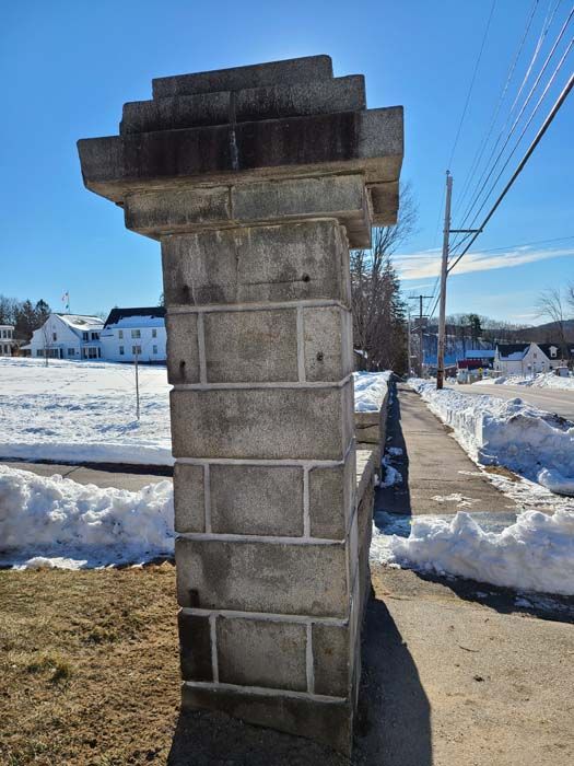 Stone pillar with decorative top, part of a low wall along a sidewalk on a sunny day with snow.