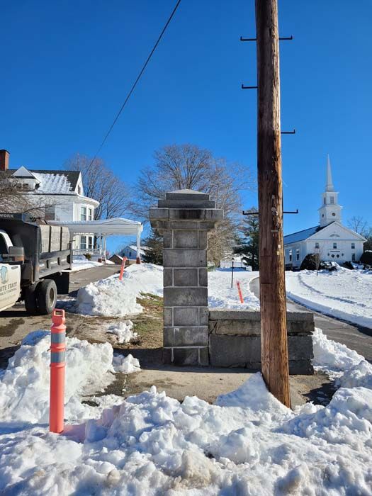 Snowy street scene: stone column, utility pole, church, white house, and truck under a blue sky.