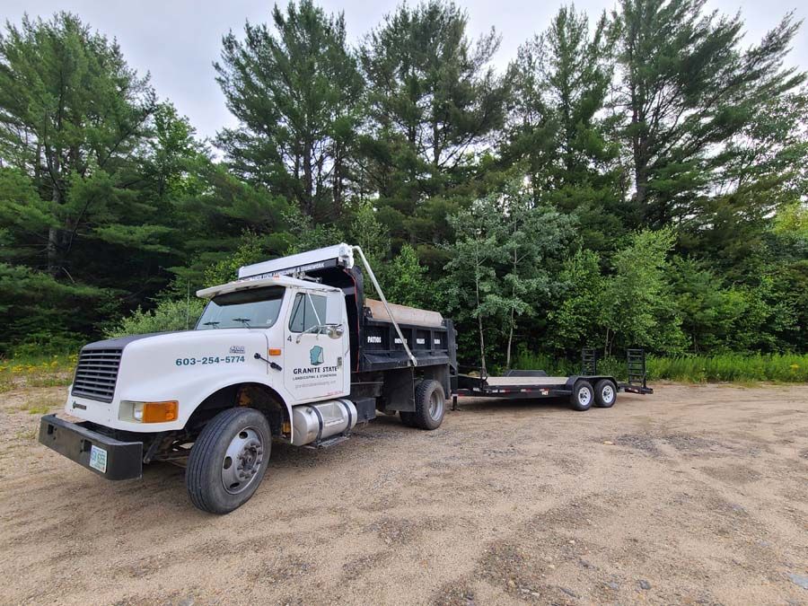 White dump truck with a flatbed trailer in a wooded area.