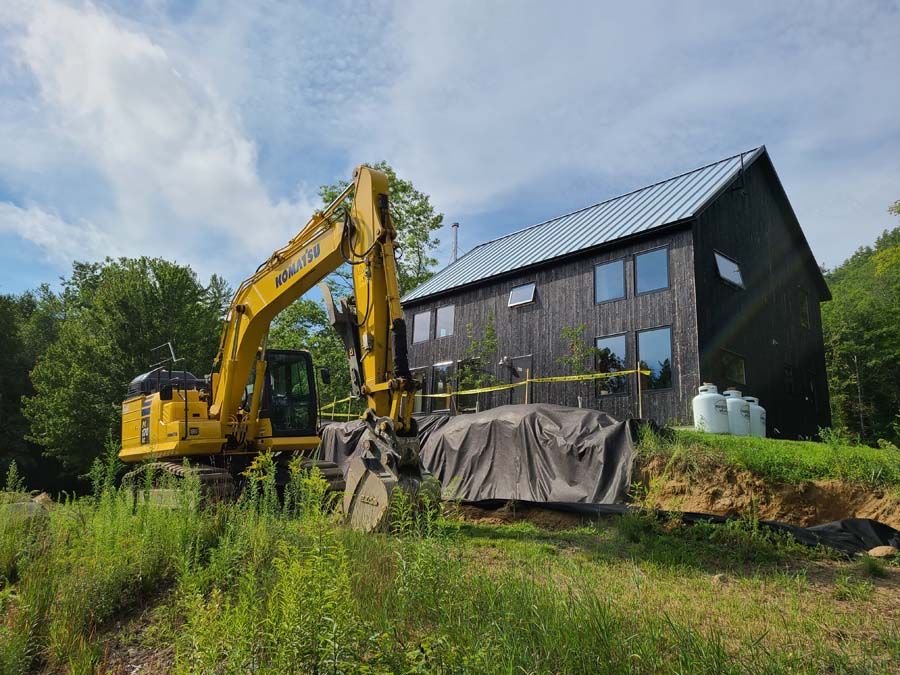 A yellow excavator sits beside a black wooden building under a blue sky.