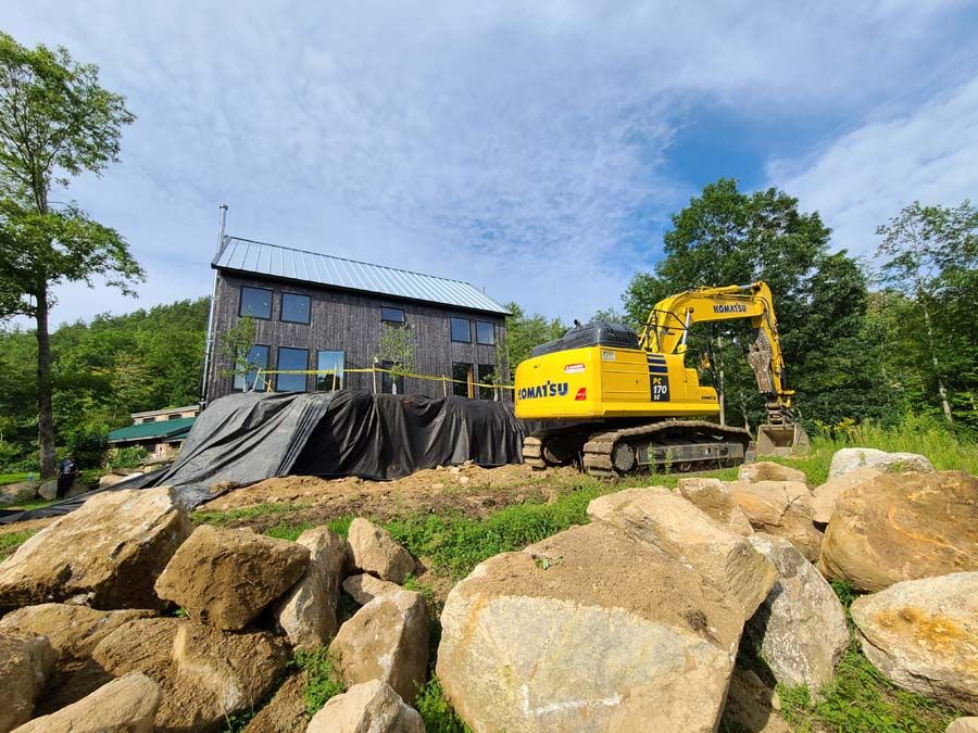 Yellow excavator next to a dark-colored house, in a wooded area, with large rocks in the foreground.