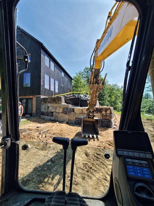 View from excavator cab toward a building with a dark facade; excavator arm and bucket over stone blocks.