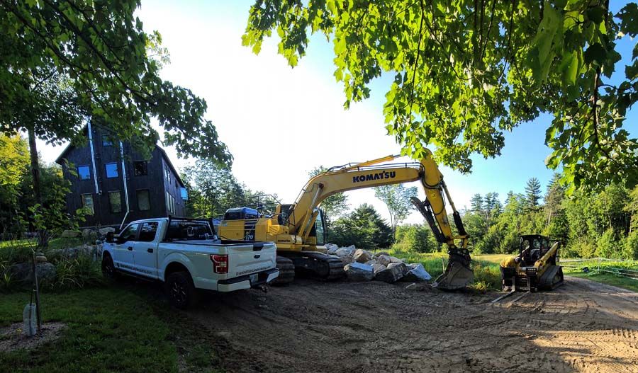 A construction site with a yellow excavator, a white truck, and a dark-colored building in the background.