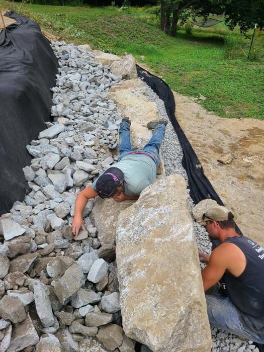 Two men building a stone wall. One is lying down, the other is standing, both working with large rocks.