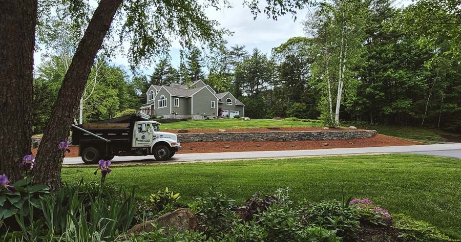 Truck delivering mulch in front of a house with a stone wall and trees on a sunny day.