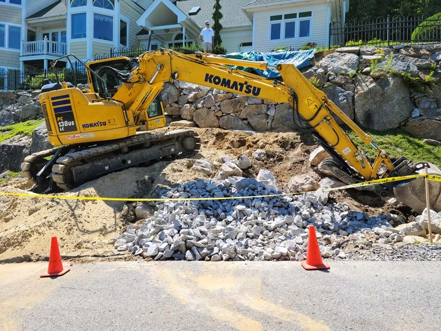 Yellow excavator on a hillside, breaking up rocks. Road in the foreground, houses in the background.