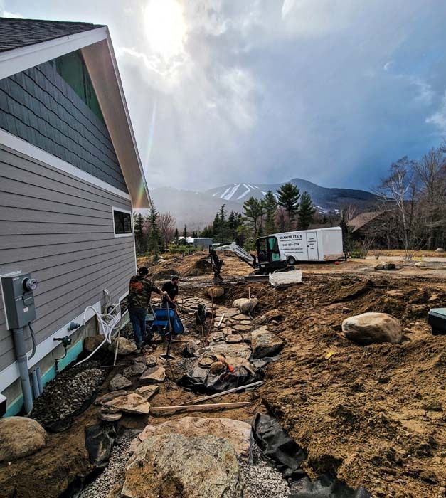 Construction site with workers, rocks, and a building. Mountain backdrop under cloudy sky.