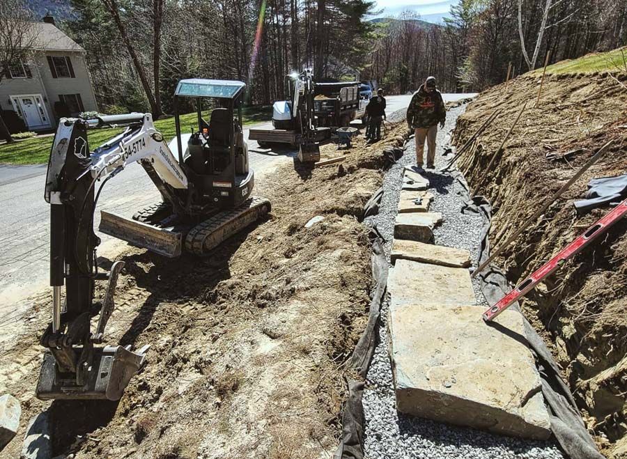 Construction site. Excavator digging alongside a stone wall under construction on a roadside. Workers present.