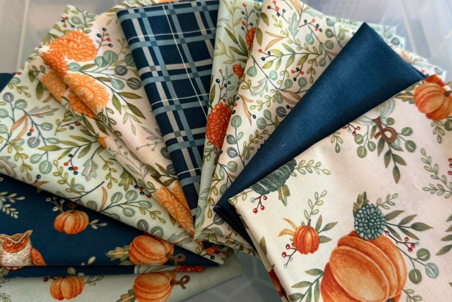 Pile of folded autumn-themed fabrics featuring pumpkins, plaid, and foliage in shades of orange, teal, and cream.