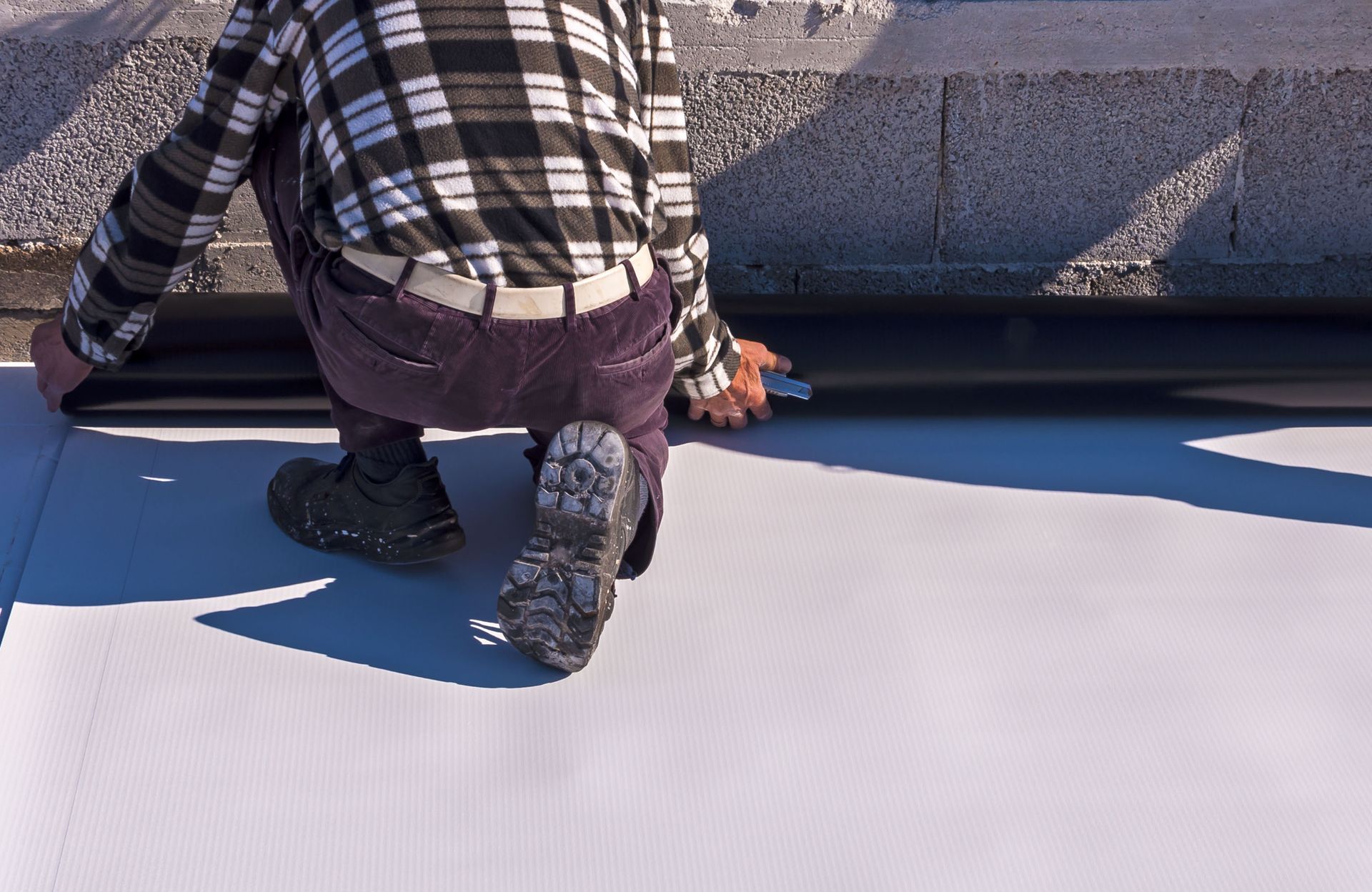Person kneeling, installing black sealant on white roofing. They wear a plaid shirt and purple pants.