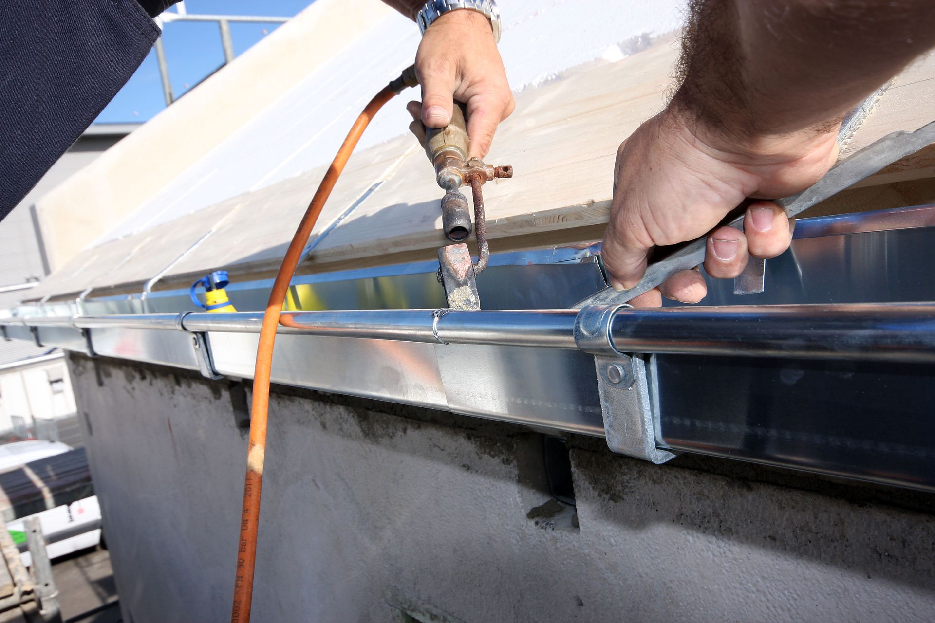 Person soldering a copper pipe to a gutter on a building's roof, outdoors, sunny.