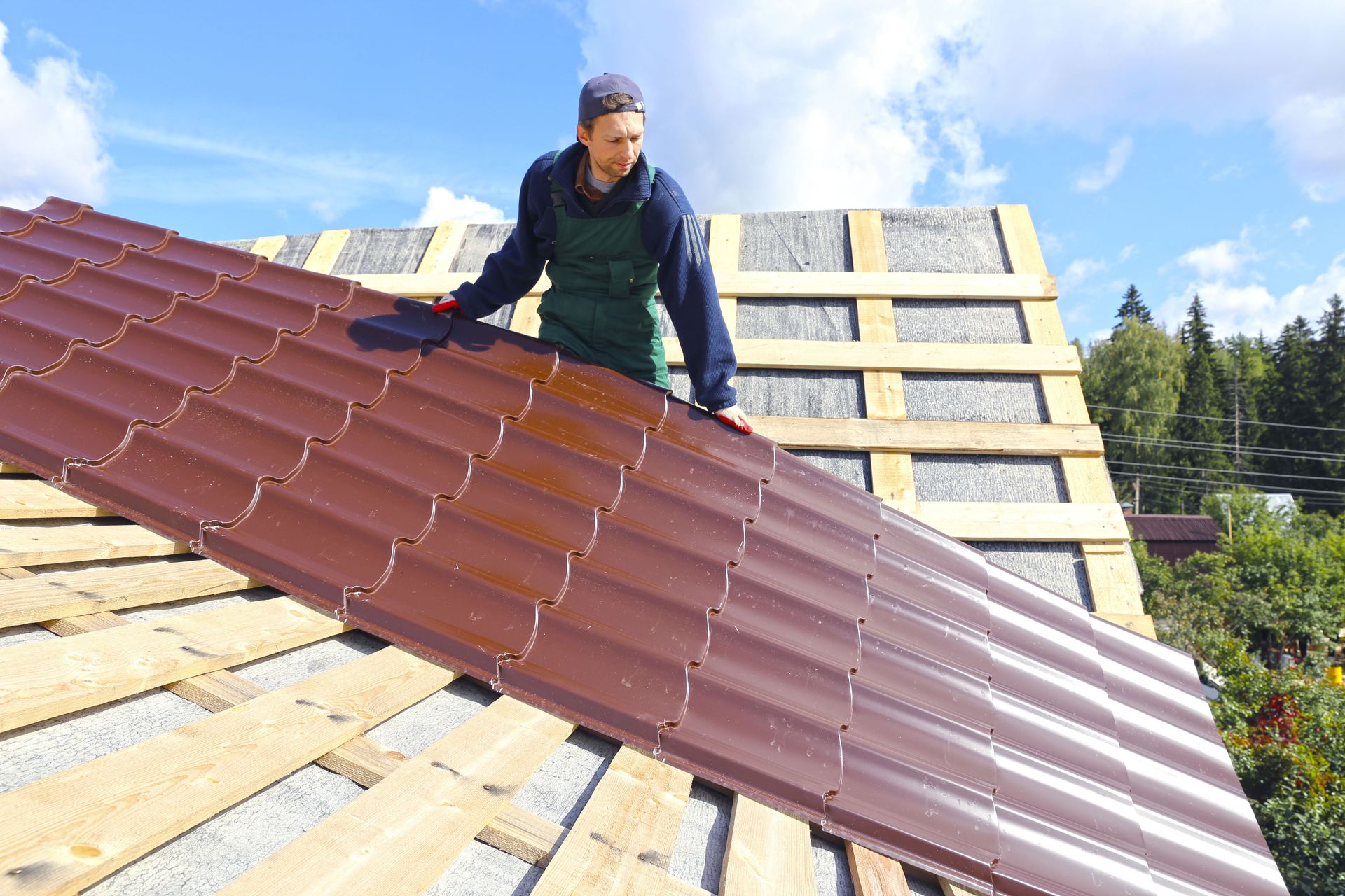 Man on a roof installing brown metal roofing panels on a sunny day.