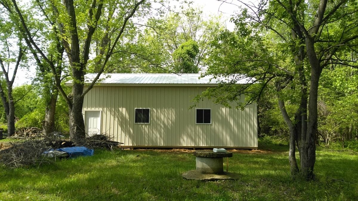 Small beige shed in a wooded yard, surrounded by green trees and grass.