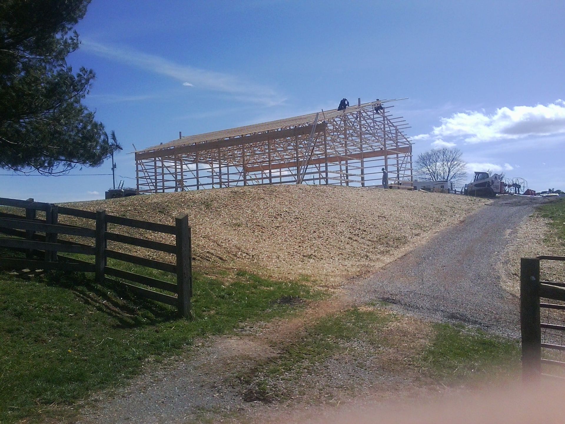 Partially built wooden barn on a hill beside a gravel driveway under a blue sky
