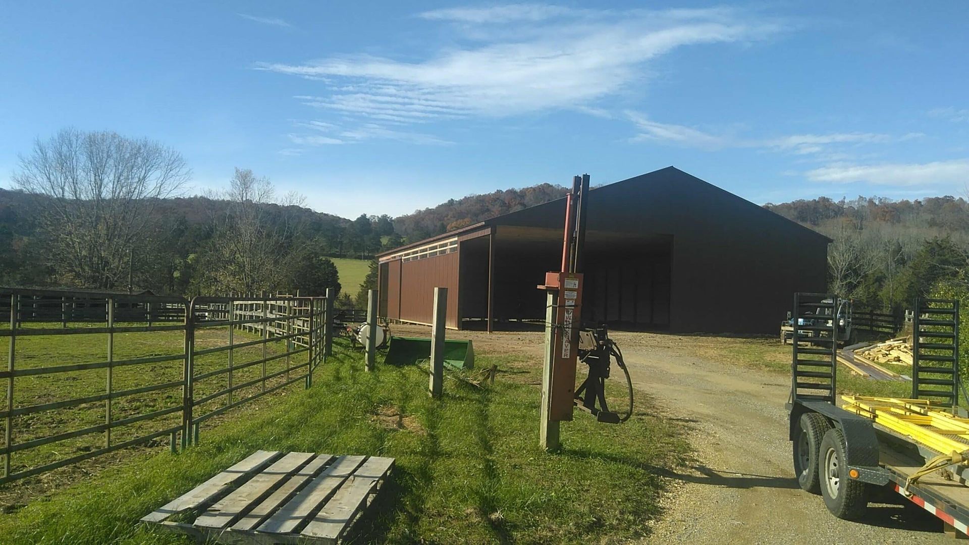 Rural barn and fenced pasture under a clear sky, with farm equipment and a wooden pallet in the foreground.