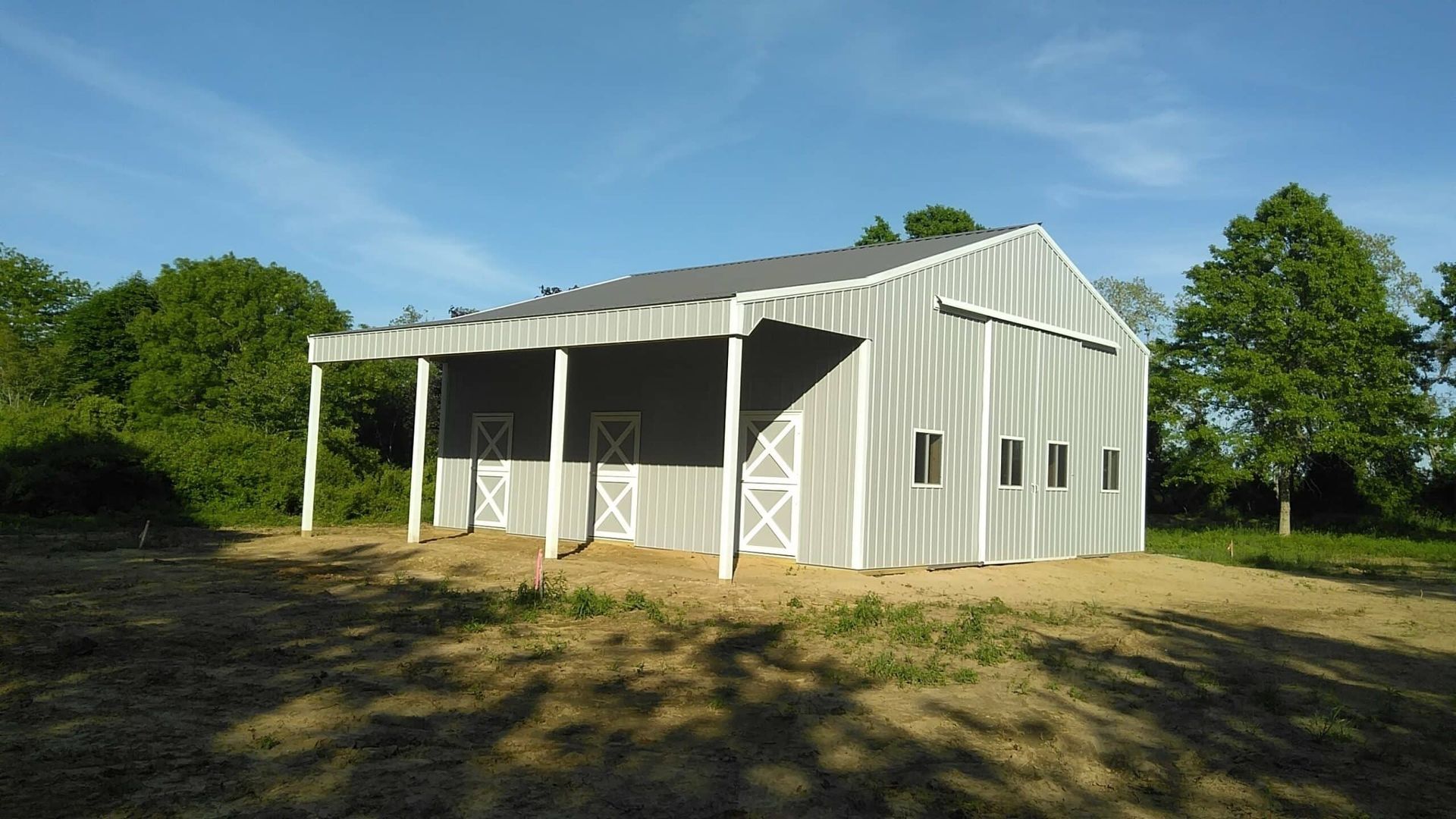White metal barn with open front doors and side windows on a grassy lot, surrounded by trees