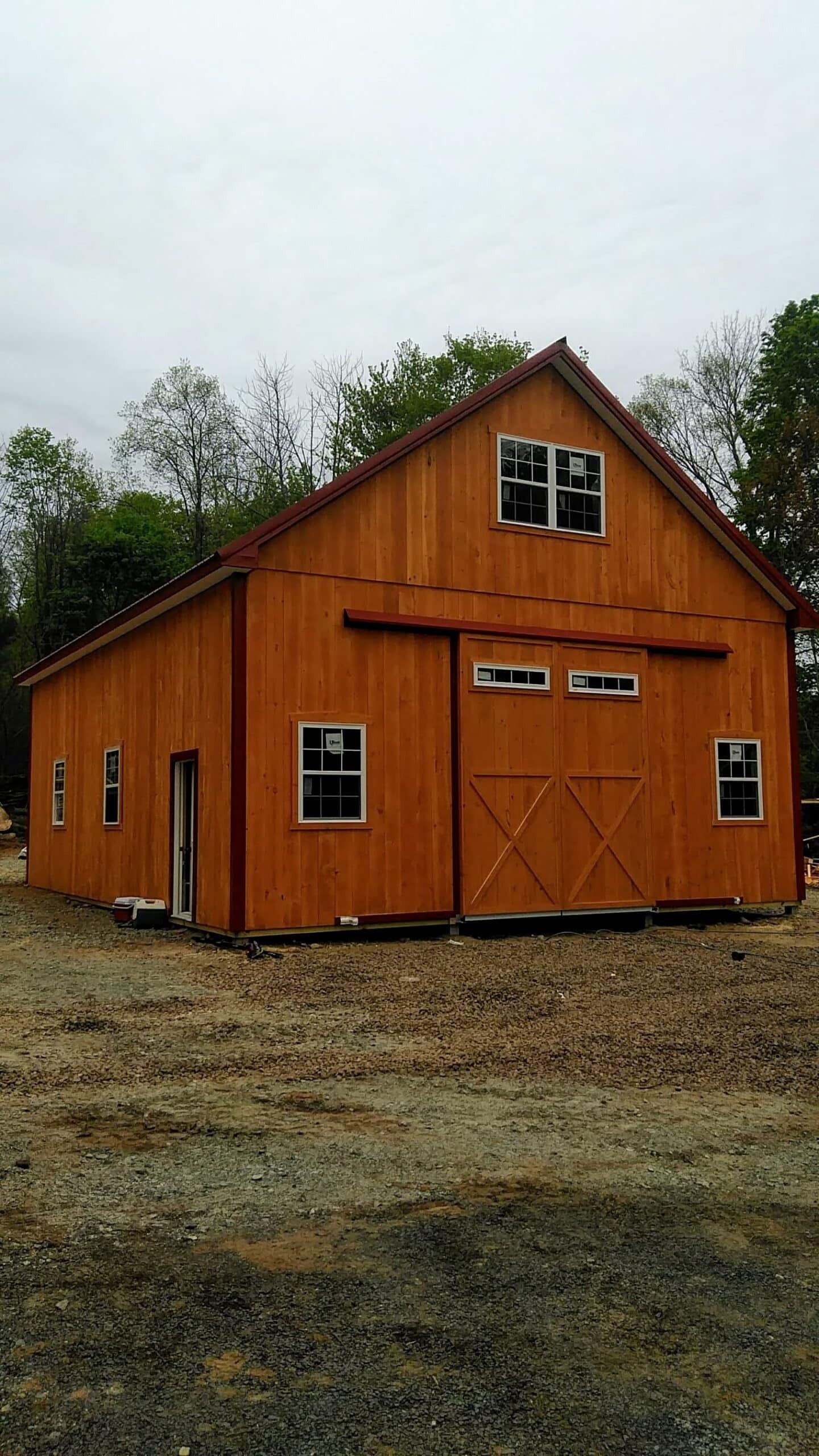 Orange wooden barn with a gambrel roof and double doors on a gravel lot under a cloudy sky