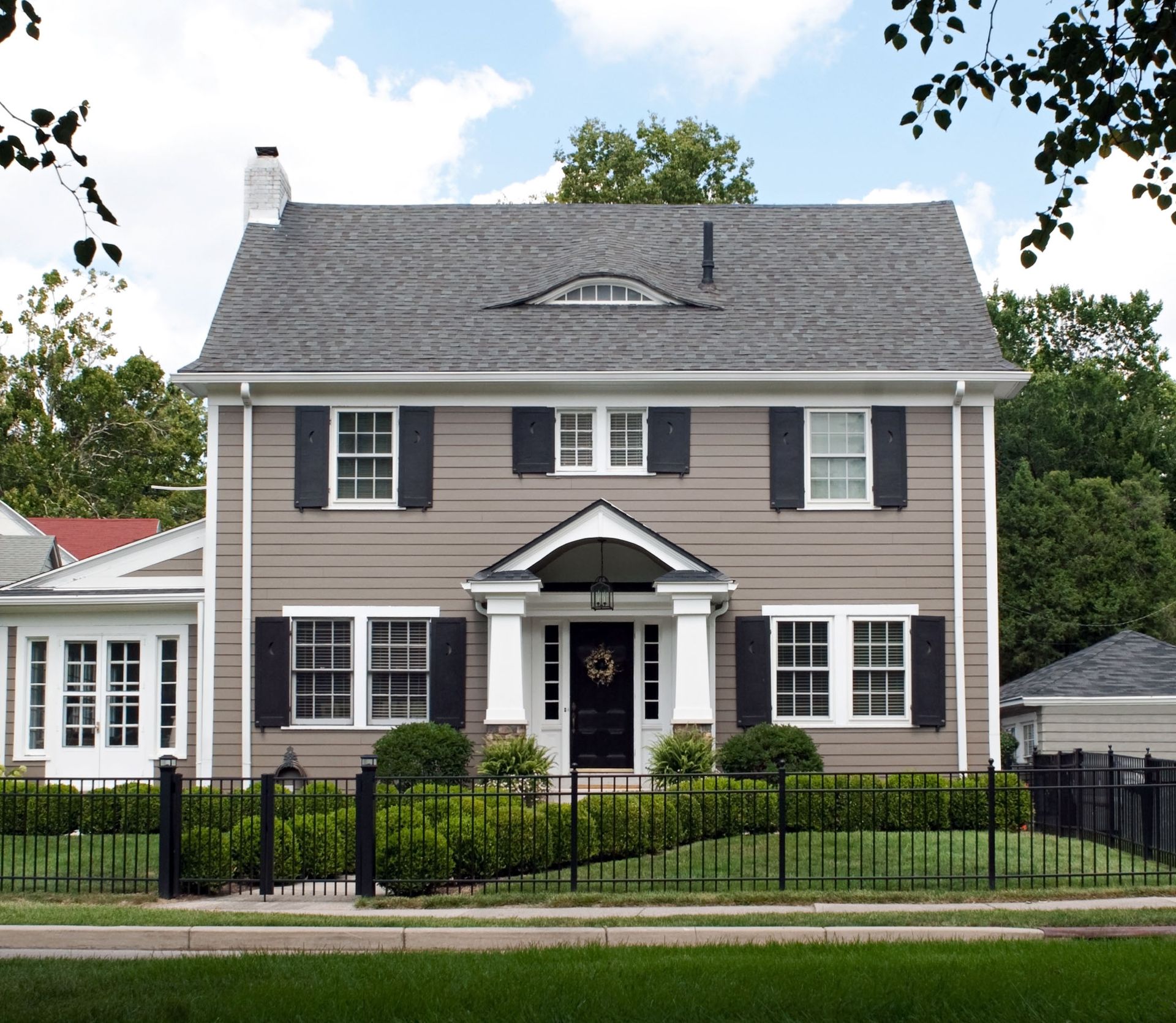Two-story gray house with black shutters and a white porch, behind a black iron fence and trees