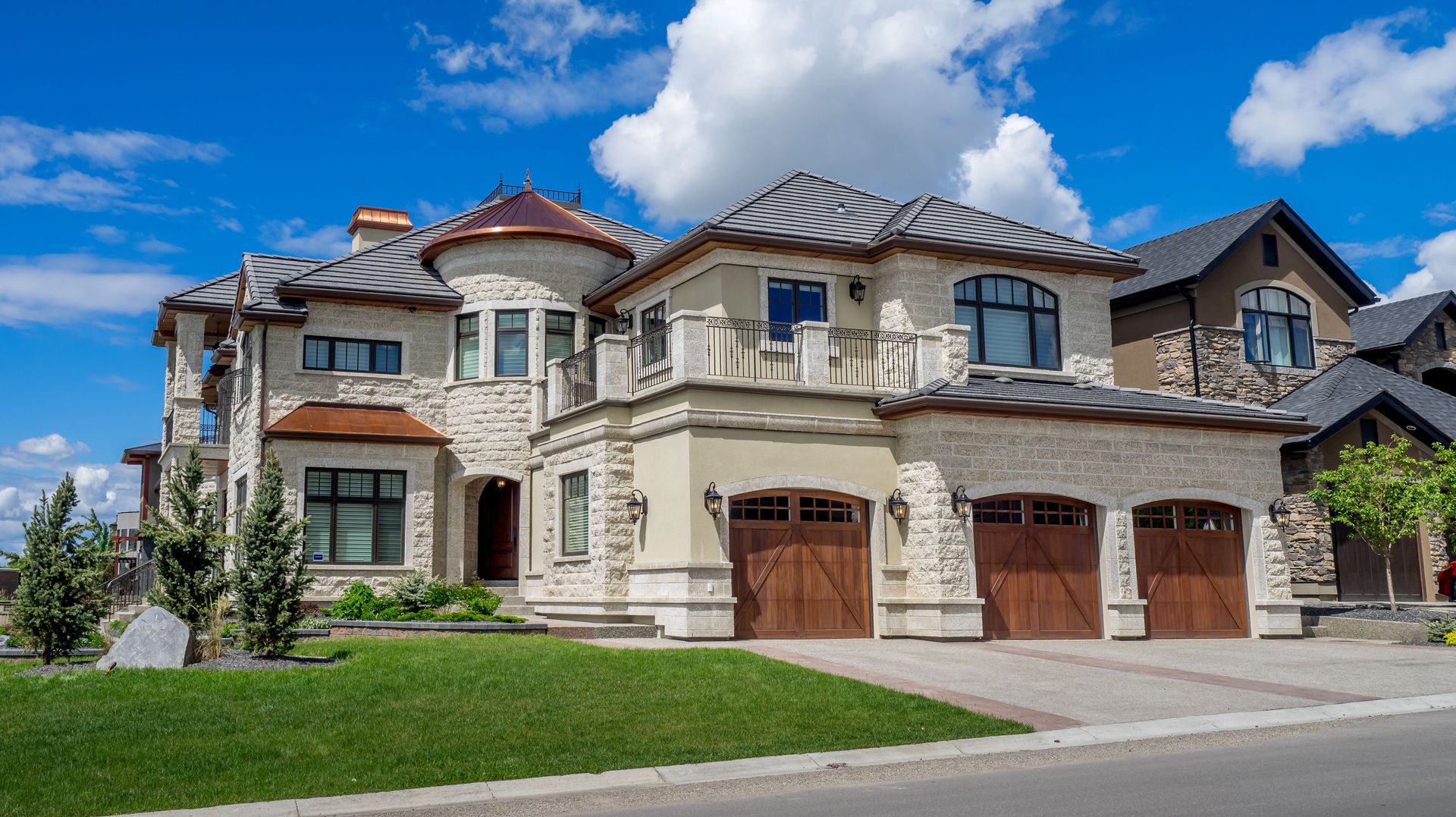 Large stone suburban house with a three-car garage, red-tiled roof, and manicured lawn under a blue sky