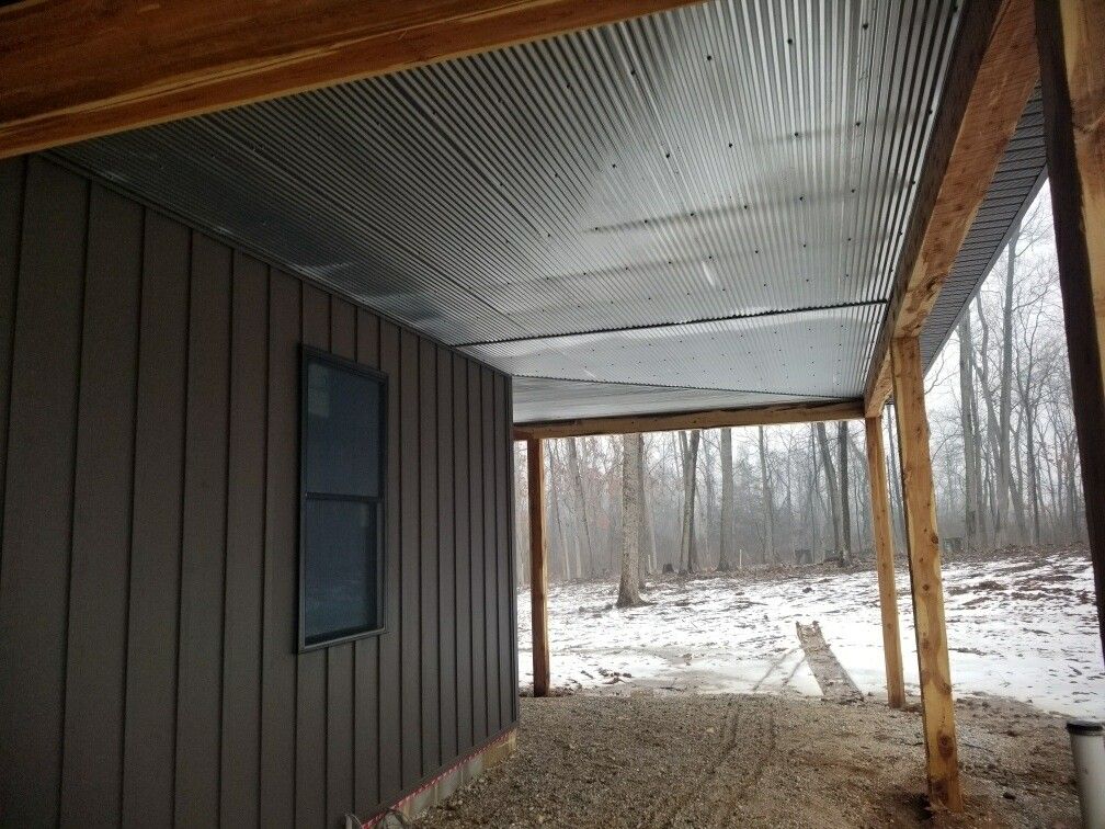 Covered porch with brown siding, metal roof, and snowy woods beyond.