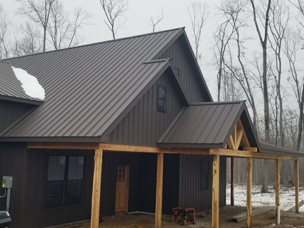 Dark metal-roofed cabin with a covered porch, set in a snowy wooded area