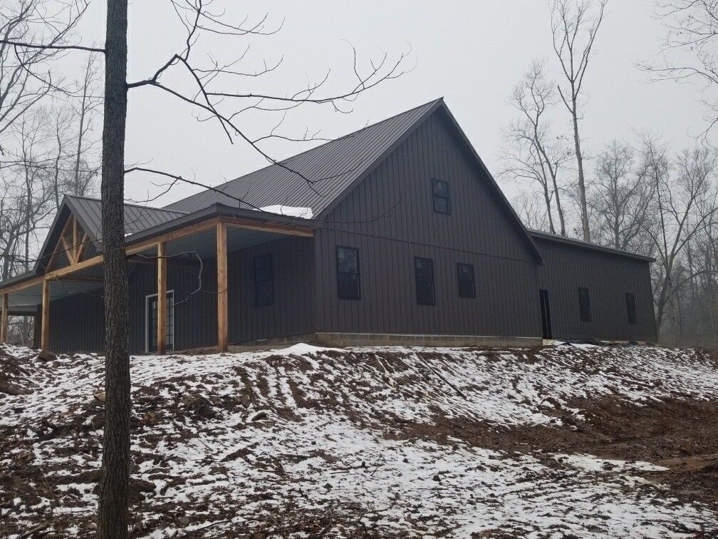 Dark gray cabin on a snowy hillside with bare trees under an overcast sky