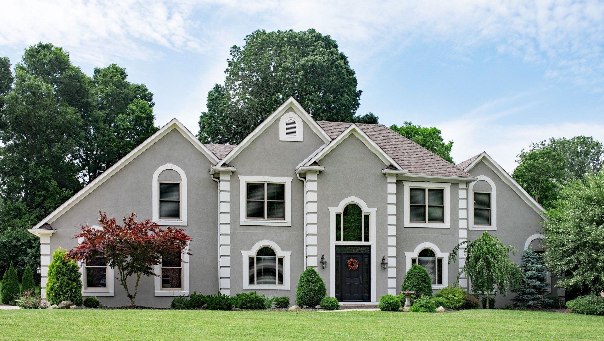 Large gray suburban house with white trim, arched windows, and a manicured green lawn under a blue sky