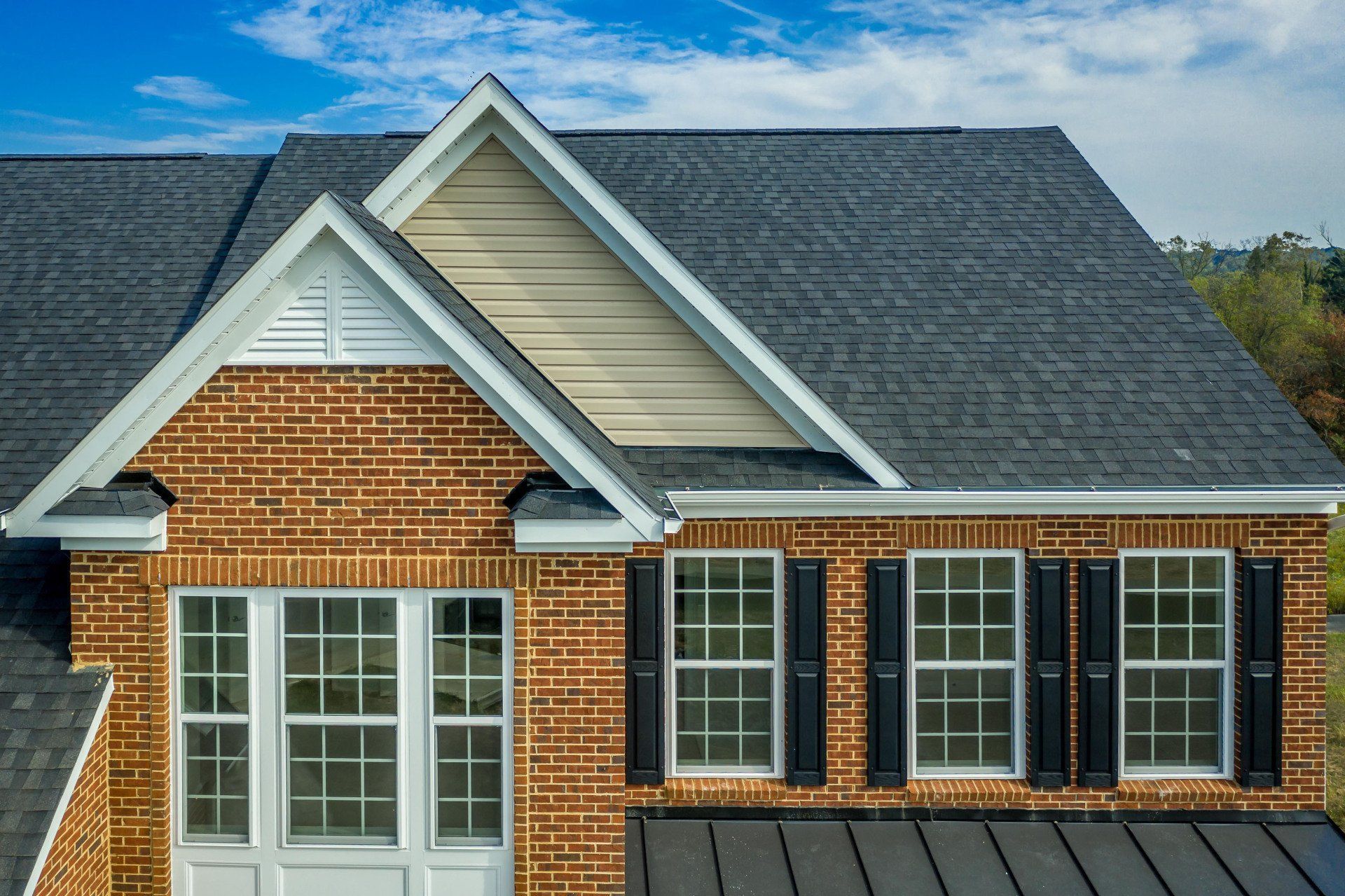 Brick suburban house with dark gray shingle roof and white-trimmed windows under a blue sky