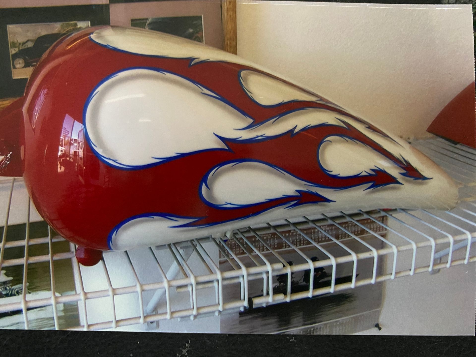 Red and white motorcycle gas tank with flame design, blue accents, sitting on a wire shelf.