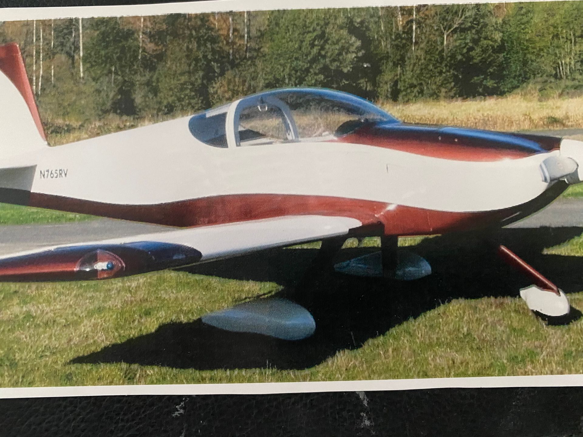 White and maroon single-engine airplane parked on grass near a runway with trees in the background.