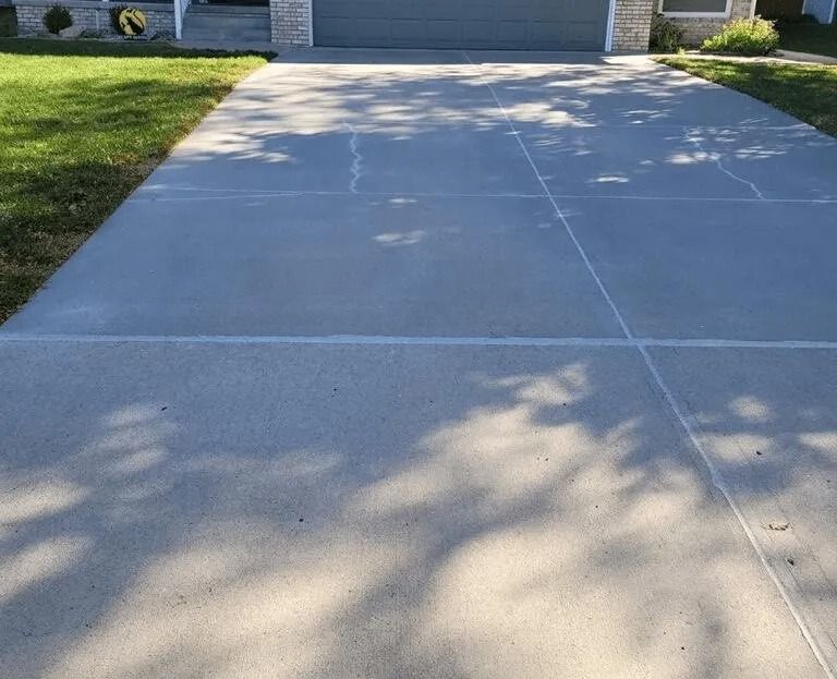 Gray concrete driveway in front of a house, with cracks and tree shadows.