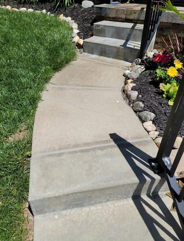 Concrete walkway leading to concrete steps, bordered by grass and flowerbed.