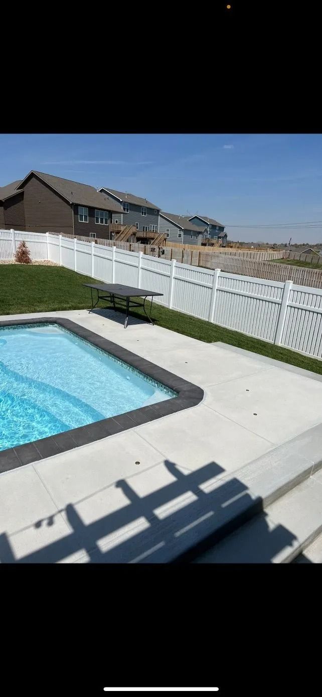 A rectangular pool with a white fence, houses in the background. Sunny day.