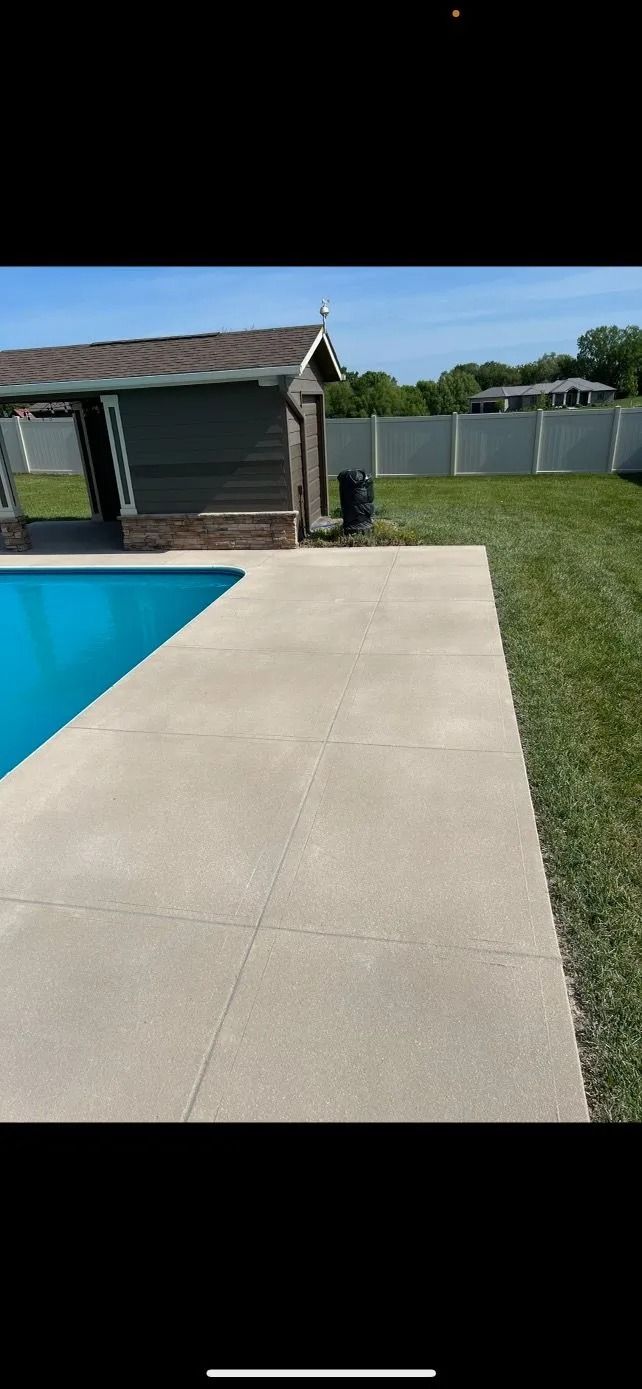 A backyard with a pool, concrete patio, shed, grass, and a white fence on a sunny day.