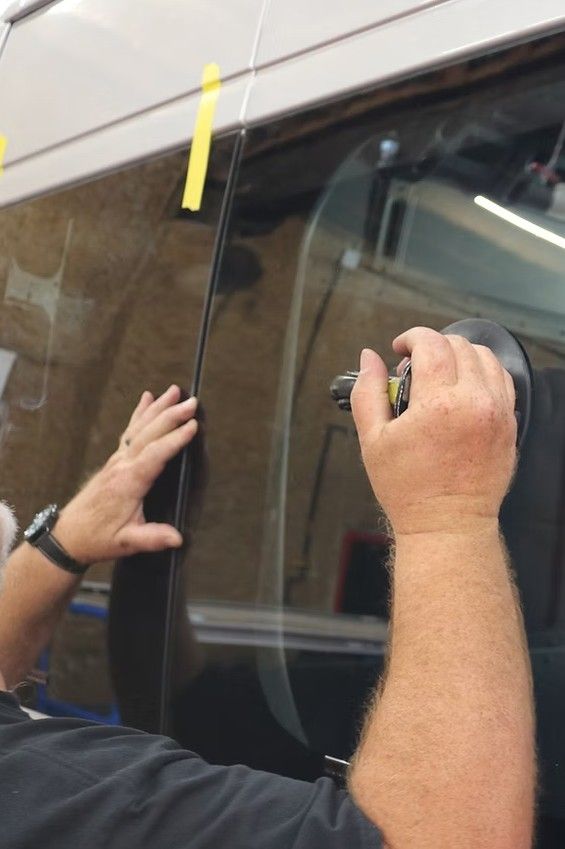 Hands applying film to a dark-tinted vehicle window with a tool and yellow tape.