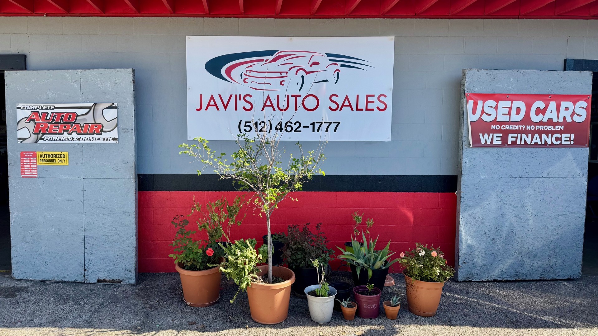 Used car lot with various cars parked outside a single-story building; flags read USED CARS.