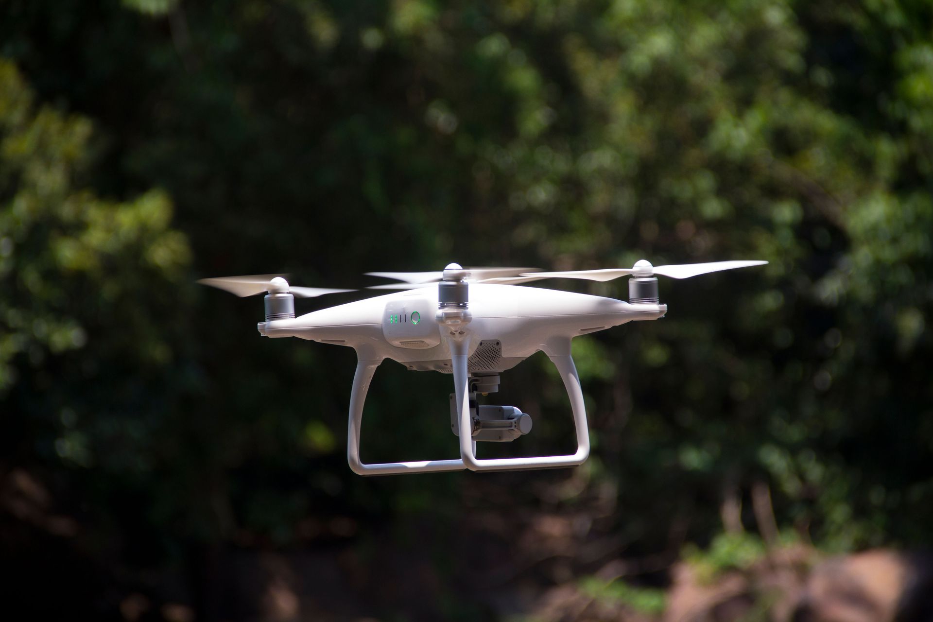 A white drone is flying in the air with trees in the background.