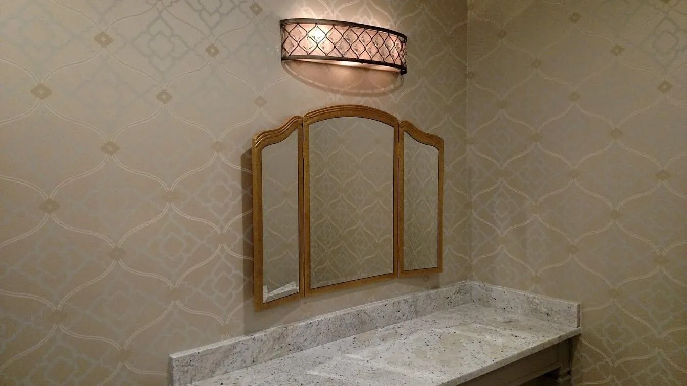 Vanity area with a trifold mirror, granite countertop, and patterned wallpaper. A light fixture is mounted above the mirror.