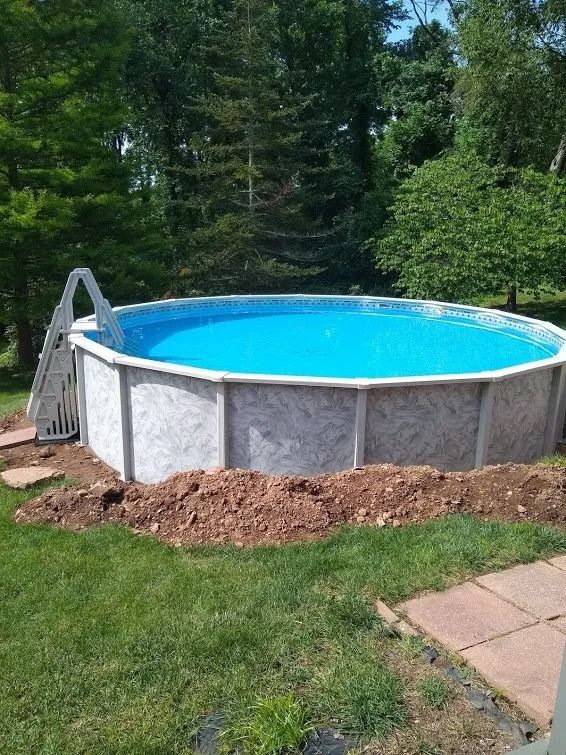 Above-ground pool with blue water surrounded by dirt and green grass, trees in the background.