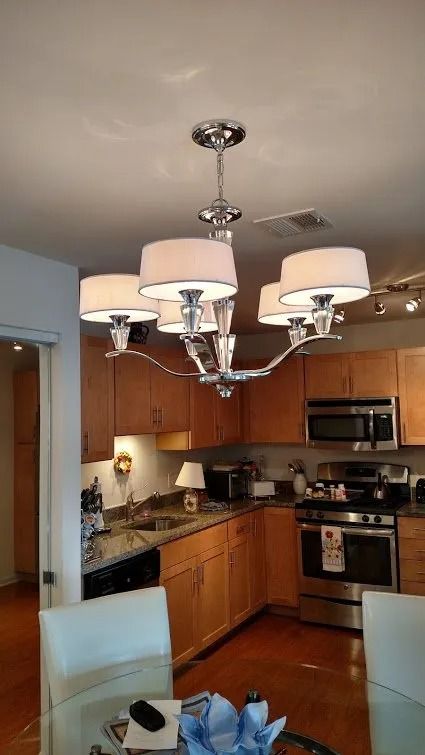 Chrome chandelier with five white lampshades over a kitchen with wood cabinets and stainless steel appliances.