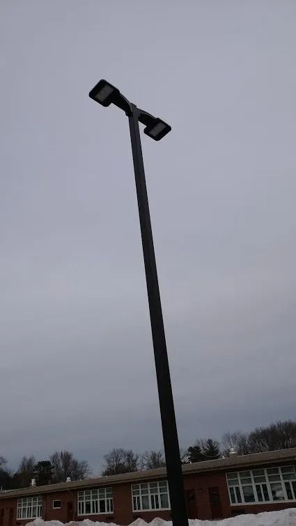 Black light pole with two rectangular lights against a cloudy sky. Brick building in the background.
