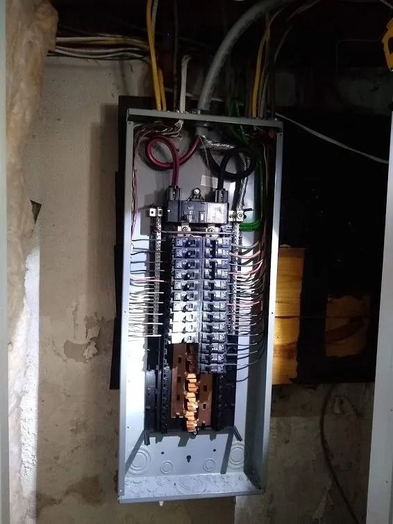 Open electrical panel with numerous circuit breakers and wiring in a basement.