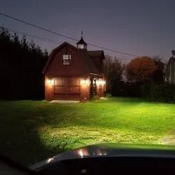 Brown barn lit up at night with green lawn, power lines, and a car headlight.