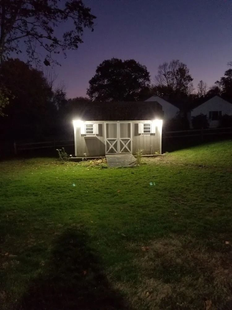 Shed in backyard lit by bright lights at dusk. Grassy lawn with trees and houses in the background.