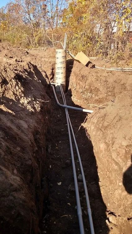 Trench with buried pipes leading to a cylindrical structure in a yard. Soil is brown, sky blue.